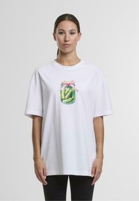 Žena s hnědými vlasy, která nosí bílý oversized tričko s ilustrací sklenice s okurkami uvázanou růžovou stuhou.