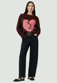 Pulôver de malha burgundy com um design de coração partido rosa, combinado com jeans escuros de cintura alta e sandálias pretas planas. Tecido texturizado.