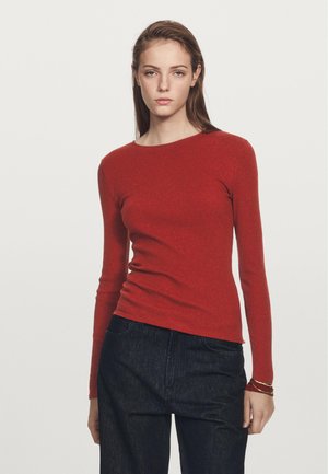 Haut rouge à manches longues en côte avec un col rond. Le tissu semble doux et ajusté. Associé à un jean bleu foncé et des bracelets superposés.