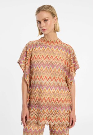 Vrouw draagt een losse top en bijpassende broek met een meerkleurig zigzagpatroon, staand tegen een effen witte achtergrond.