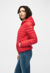 Γυναίκα με κυματιστά μαλλιά, που φοράει κόκκινο μπουφάν με κουκούλα και μπλε τζιν, όρθια πλαγίως με το ένα χέρι στην τσέπη του μπουφάν.