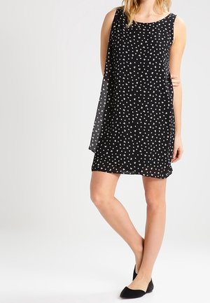 Robe noire sans manches avec un motif à pois de fleurs blanches, tissu doux et un voile fluide qui tombe d'un côté.