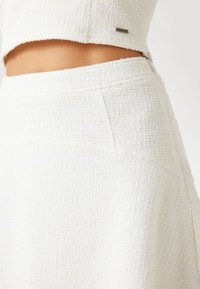 Valge tekstuuriga kangast komplekt, mis sisaldab lühikest topi ja seelikut, millel on sile vöökoht ja väike hõbedane detail topil.
