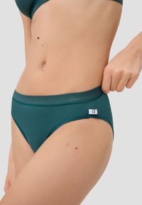Dunkelgrüne Damenunterwäsche aus glattem Stoff, mit einem kontrastierenden Taillenbund und einem Etikett mit einem Herzsymbol an der Seite.