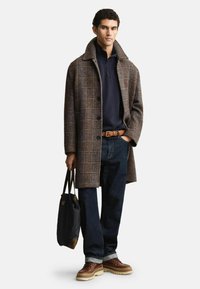 Cappotto lungo a quadri marrone con colletto alto, polo blu navy, jeans scuri, scarpe di pelle marrone e borsa tote nera.