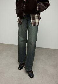 Getailleerde bruine jas van nep bont over een geruite blouse met knopen, gecombineerd met wijde blauwe spijkerbroeken en zwarte loafers met gouden details.