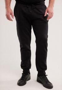 Mees mustade jooksupükste ja mustade paeltega spordijalatsitega seisab ühevärvilise tausta ees.