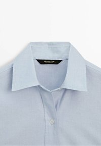 Chemise bleu clair avec col et patte de boutonnage, dotée d’un col pointu classique et d’une étiquette de marque visible à l’intérieur de l’encolure.
