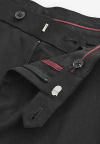 Schwarze Hosen mit glatter Textur, ausgestattet mit einem Knopfverschluss, einem Reißverschluss und roten Akzenten am Bund. Enthält ein kleines silbernes Detail.