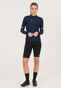 Maglia da ciclismo blu navy con zip frontale completa, maniche lunghe e vestibilità slim; abbinata a pantaloni corti da ciclismo neri. Presenta loghi e dettagli a contrasto.