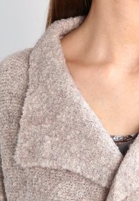 Beżowy sweter o teksturze, z dużym kołnierzem, wykonany z miękkiego, puszystego materiału, z warstwowym designem i widocznymi detalami przeszycia.