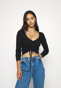 Black cropped long-sleeve top with a V-neck and gathered detail at the front, paired with high-waisted blue jeans.