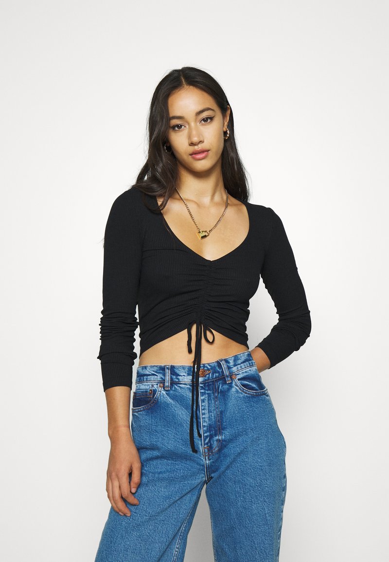 Black cropped long-sleeve top with a V-neck and gathered detail at the front, paired with high-waisted blue jeans.