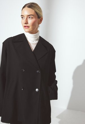 Blonde woman with slicked-back hair wearing a white turtleneck and oversized black double-breasted coat, looking left against a white background.