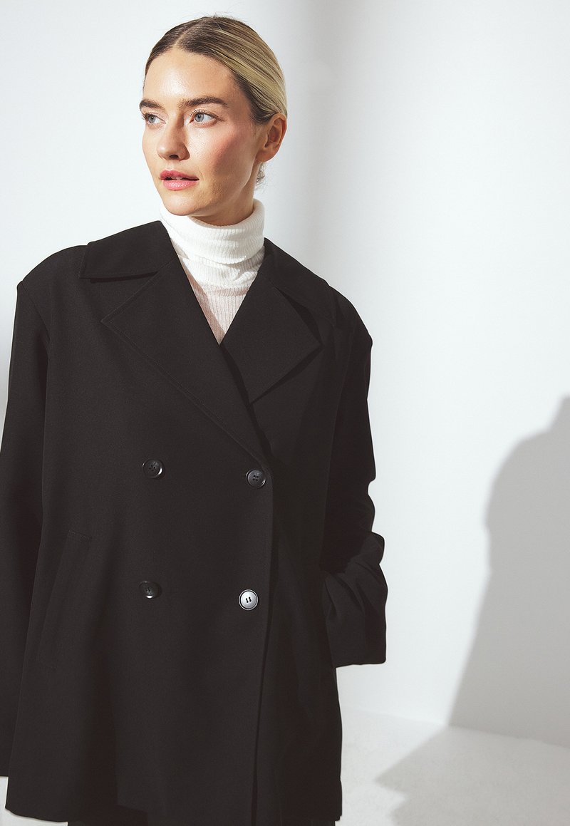 Femme blonde aux cheveux plaqués en arrière portant un col roulé blanc et un manteau noir oversize à double boutonnage, regardant à gauche sur fond blanc.
