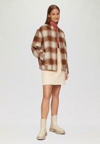 Bruine en crèmekleurige geblokte jas met een hoge kraag, gecombineerd met een lichtbeige rok en crèmekleurige enkellaarsjes. Glad textuur in het geheel.