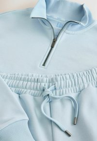 Pulover albastru deschis cu fermoar pe jumătate, guler cu nervuri și tiv cu șnur. Material moale cu o textură gathered la talie. Accesoriu cu fermoar argintiu.