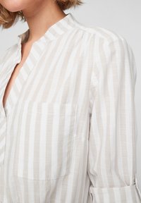 Blouse rayée beige clair en tissu doux et texturé. Présente un col et une poche unique sur le côté gauche.