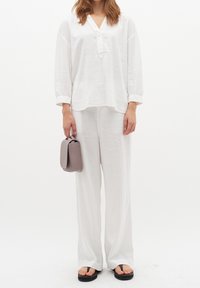 Top in lino bianco con scollatura a V e maniche lunghe, abbinato a pantaloni larghi in lino bianco. Tiene in mano una piccola borsa grigia ed indossa infradito neri.