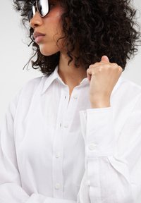 Camisa blanca de cuello, hecha de un tejido ligero, con botones en la parte delantera y un único bolsillo en el pecho; combinada con gafas de sol oscuras.