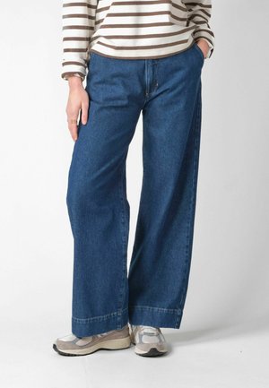 Person trägt blaue Jeans mit weitem Bein, beige-weiße Sneakers und ein beige-weiß gestreiftes Langarmshirt, die eine Hand in der Tasche hat.