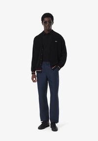 Cárdigan negro con puños y ribete acanalados, polo negro, pantalones azul marino y zapatillas negras. Los accesorios incluyen gafas de sol oscuras.