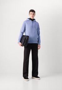 Hanorac bleu deschis cu buzunar frontal tip kangaroo, geantă de umăr neagră, pantaloni largi negri și pantofi sport negri cu talpă albă.