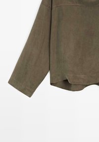 Manche de chemise longue en daim vert olive et ourlet inférieur avec couture visible sur fond blanc.