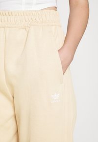 Ljust beige Adidas sweatpants med elastisk midja, sidofickor och en liten vit broderad logga på benet. Mjuk textur.