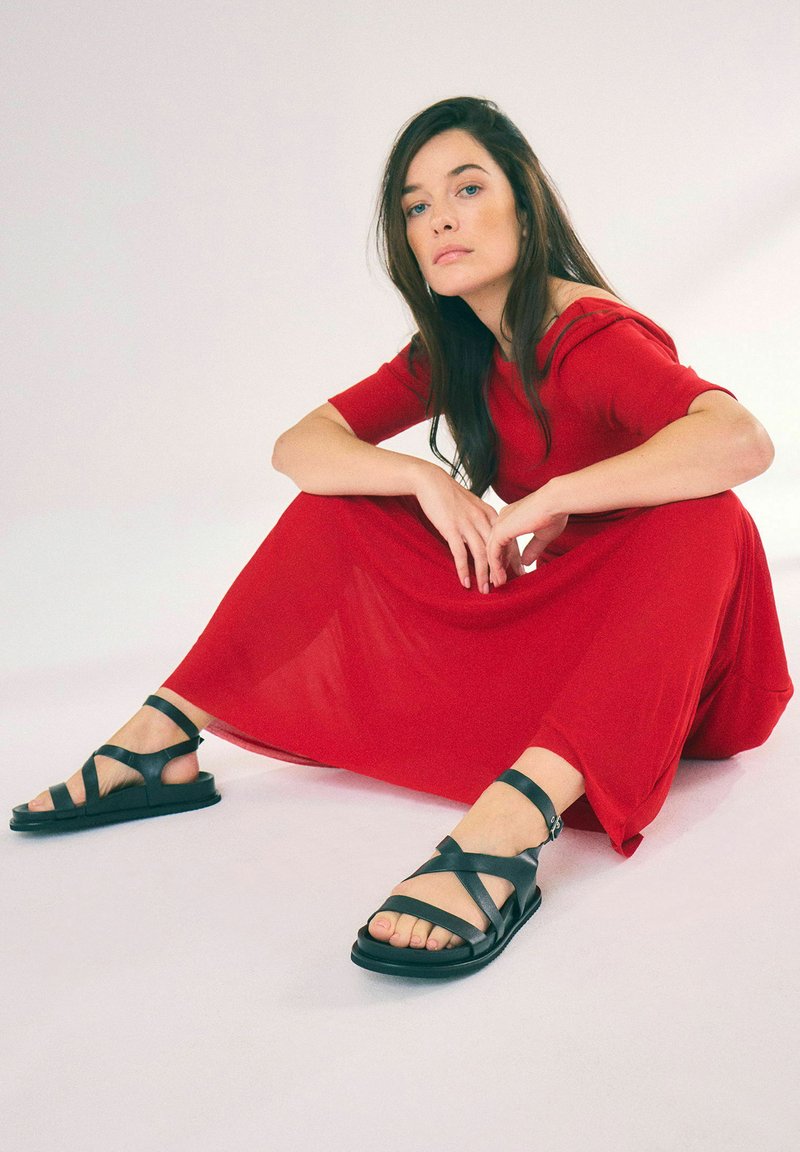 Rotes Kleid mit kurzen Ärmeln, fließender Stoff, kombiniert mit schwarzen Sandalen mit Riemen, flacher Sohle und Knöchelriemen, vor einem hellen Hintergrund.