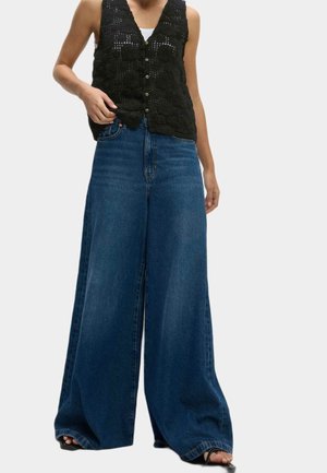 Person iført sort hæklet knappet vest over hvid top og vidbenede blå denimjeans med åbne sorte sandaler.