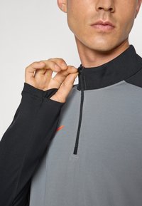 Grijze en zwarte quarter-zip top met lange mouwen; gemaakt van soepele stof. Zwarte accenten bij de kraag en manchetten; heeft een contrasterend oranje logo.