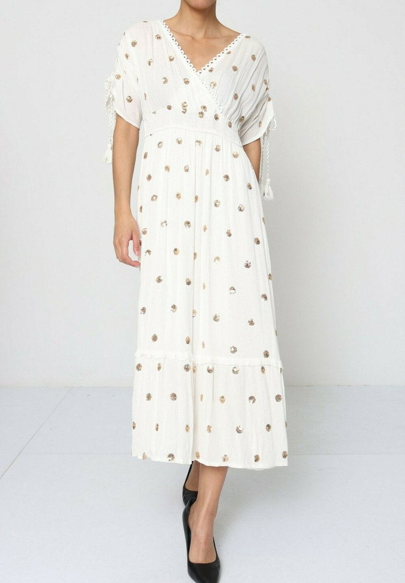 Robe midi blanche avec un design enroulé, manches courtes, tissu texturé, accents dorés à pois et franges sur les côtés. Assortie avec des talons noirs.