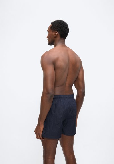 Μυώδης άνδρας με σκουρόχρωμο δέρμα στέκεται γυμνόστηθος στραμμένος προς τα αριστερά, φορώντας σκούρο μπλε σορτς, σε απλό λευκό φόντο.