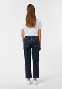 Jeans in denim blu scuro con taglio dritto, caratterizzati da cuciture a contrasto, due tasche posteriori e orli alla caviglia. Abbinati a una t-shirt bianca.