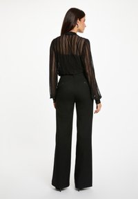 Donna vista di spalle che indossa una blusa nera trasparente con righe verticali e pantaloni larghi a vita alta neri, in piedi su uno sfondo bianco.