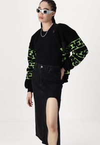 Saia de ganga preta com uma fenda lateral, combinada com um cardigan oversized texturizado em preto e verde néon e uma blusa justa preta. Óculos de sol brancos.