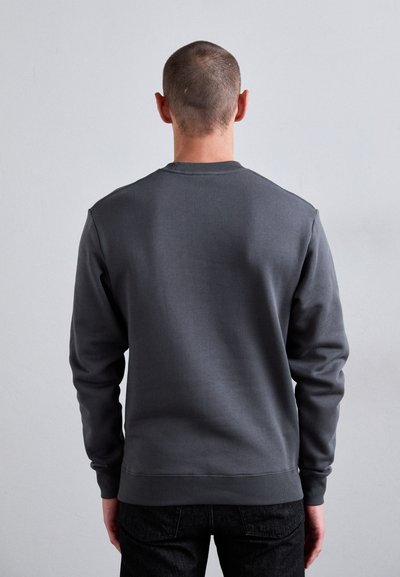 Uomo con i capelli corti che indossa una maglietta a maniche lunghe grigia scura semplice e jeans neri, di fronte a una parete grigia chiara.