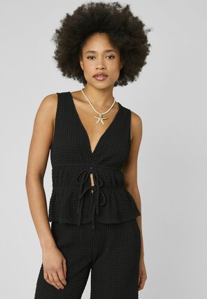 Femme aux cheveux bouclés naturels portant un haut sans manches noir texturé et un pantalon, accessoirisée d'un collier étoile de mer, debout devant un fond gris.