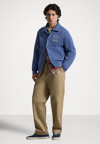 Plava pamučna jakna s okruglim izrezom i kopčanjem na gumbe, nošena preko karirane košulje. Kombinaciju upotpunjuju kaki hlače i plave tenisice.