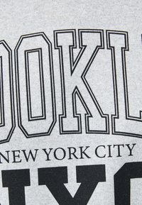 Sudadera de algodón gris con un atrevido texto negro que dice "BROOKLYN NEW YORK CITY." El texto tiene un borde grueso. Textura suave.