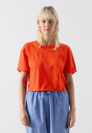 Noor inimene blondide juustega, kandes eredat oranži lühikeste varrukatega särki ja siniseid vertikaaltriibulisi pükse, taustal tavaline taust.