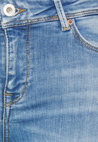 Blå denimjeans med ljus tvätt, kopparknappdetaljer, vertikal sömnad och en något sliten textur. Nära bild av tyget.