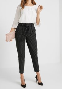 Schwarz-weiß gestreifte, maßgeschneiderte Hose mit hoher Taille und Krawatten-Detail, kombiniert mit einer weißen Bluse und einer Clutch, getragen mit schwarzen High Heels.