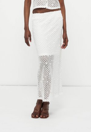 Kvinde iført en hvid hækle maxi nederdel med et ensfarvet for der ender over knæene og brune sandaler med stropper.