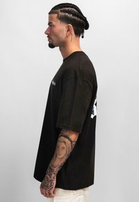 Schwarzes, übergroßes T-Shirt aus Baumwolle mit kurzen Ärmeln, das auf der Vorderseite Text und auf der Rückseite eine blaue Grafik zeigt. Das Model hat komplizierte Tattoos.