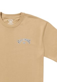 Camiseta de algodón beige con cuello redondo, mangas cortas y un pequeño logo bordado azul en el lado izquierdo. Textura suave.
