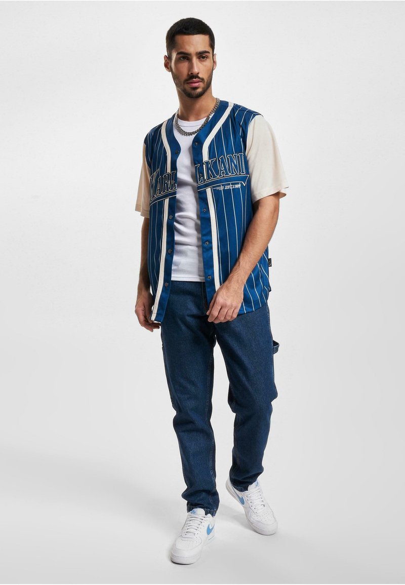 Blue and white pinstriped sleeveless jersey over a white T-shirt, paired with dark denim pants and white sneakers. Silver chain necklace visible.