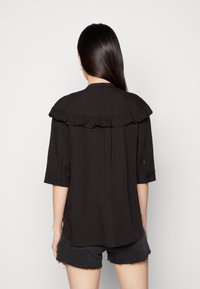 Blusa preta texturizada com gola franzida e mangas três quartos, apresentando um corte solto e fendas laterais. Usada com calções escuros.