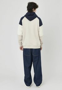 Sweat à capuche crème et marine, présentant un design coloré, des poignets côtelés et une coupe décontractée. Associé à un jean en denim foncé ample.